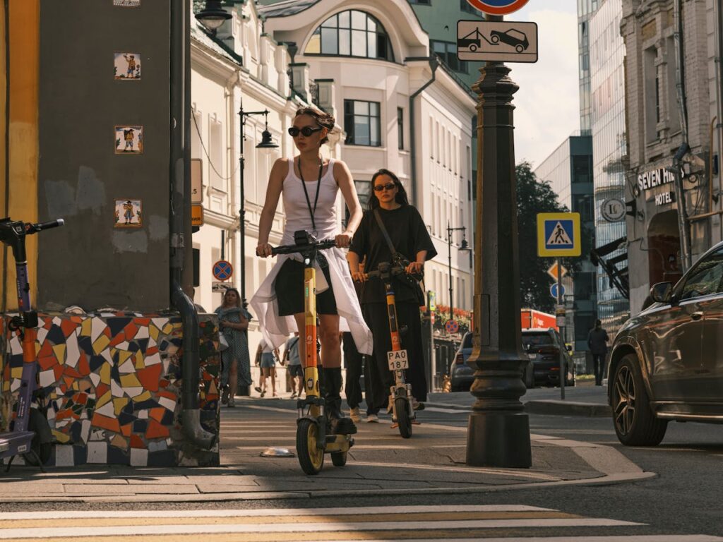 pexels-photo-26708106 Two women riding electric scooters on a city street, showcasing urban commute.